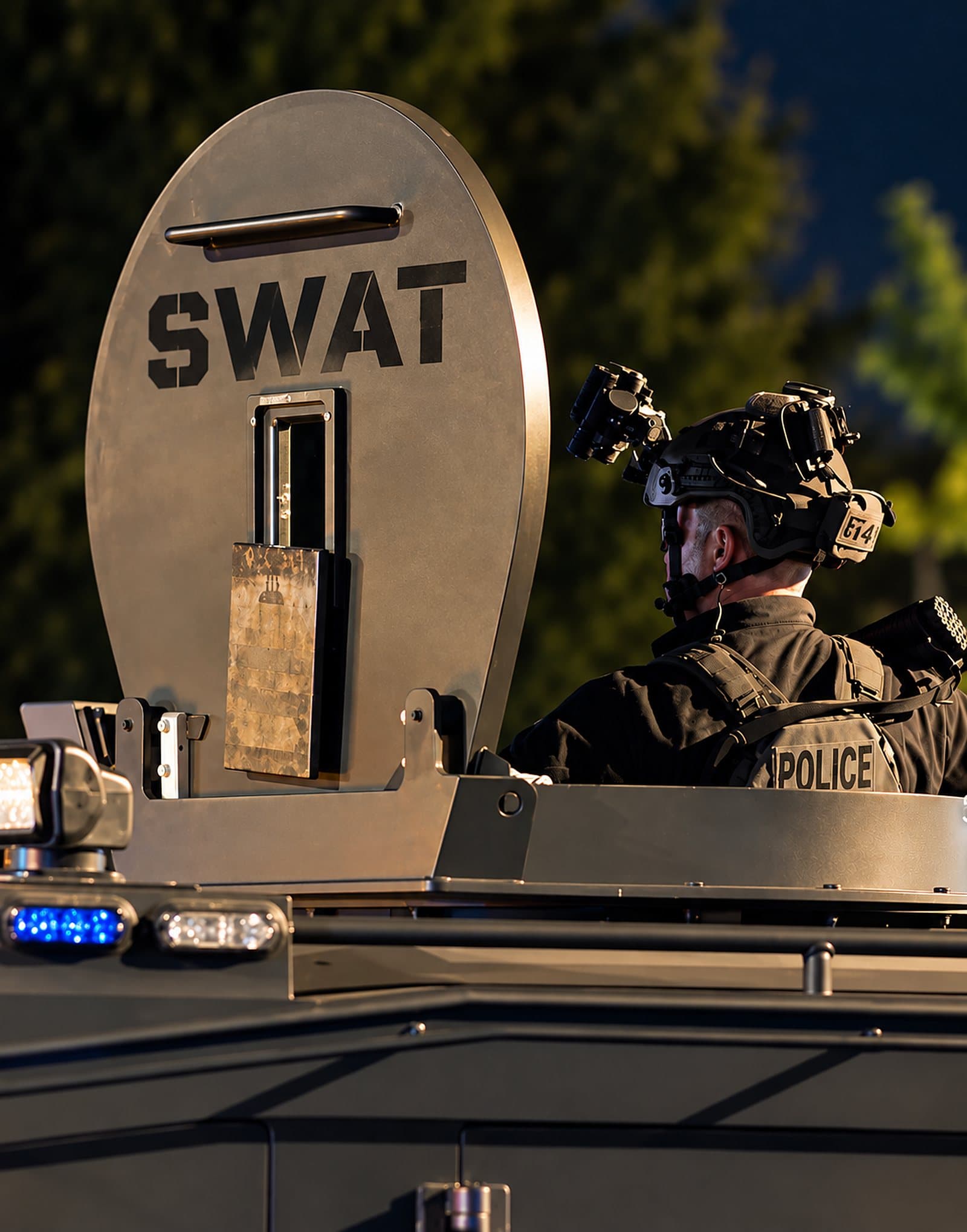 A SWAT officer behind the shield of an armored vehicle at dusk